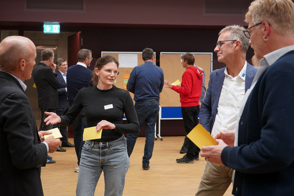 Im Vordergrund steht eine Gruppe von vier Menschen im Halbkrei, im Hintergrund steht eine weitere Gruppe vor Pinnwänden. Die Menschen sind zwischen 30 und 50 Jahre alt, tragen normale, gepflegte Kleider oder Sakko und unterhalten sich angeregt. Einige haben gelbe Zettel in den Händen.