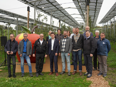 Vor einer Agri-PV-Anlage stehen rund 12 Menschen in relativ schicker Kleidung. Im Hintergrund ist ein grosser roter Apfel zu sehen. Alle sind zwischen 30 und 55 Jahre alt. Der Himmel ist grau.