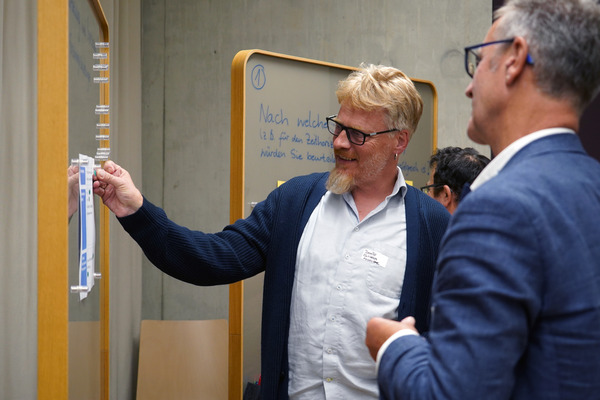 Zwei Männer im mittleren Alter stehen vor einer Stehwand mit gläserner Oberfläche. Einer hat gerade die Hand an einem Zettel auf der Wand. Beide lächeln und wirken sympathisch und angeregt.